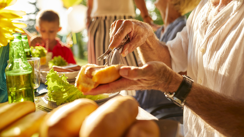 A person places a hot dog in a bun at a gathering, near a table with other people.