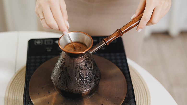 a pair of hands making Turkish coffee on a stovetop with a cezve