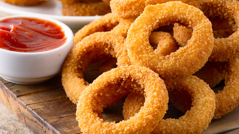 A pile of onion rings on a wooden platter next to a small bowl of ketchup.