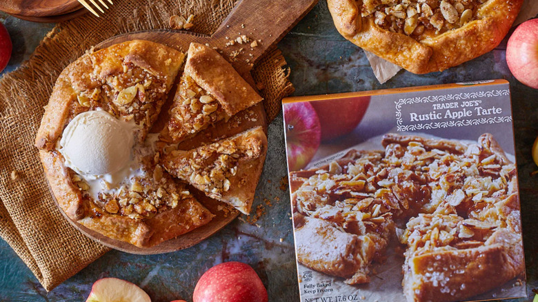 A kitchen table with a Rustic Apple Tart package next to a cooked version surrounded by apples
