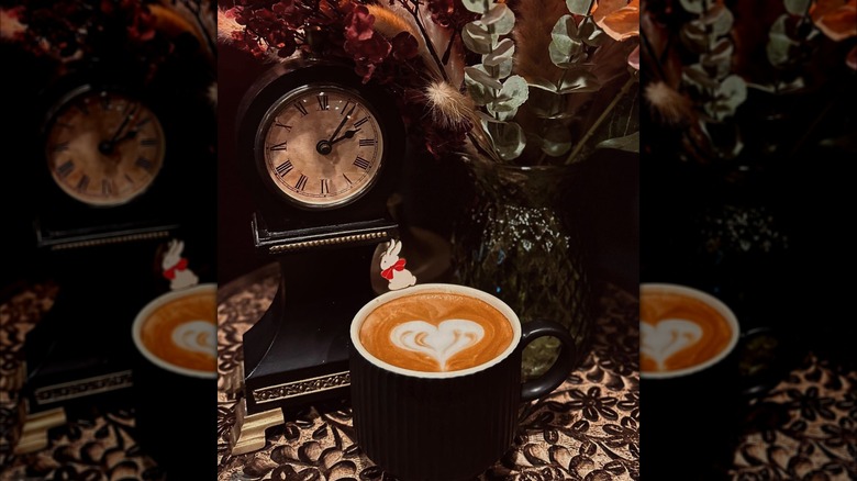 Mug with heart-shaped latte art pictured in front of decorative clock and floral arrangement