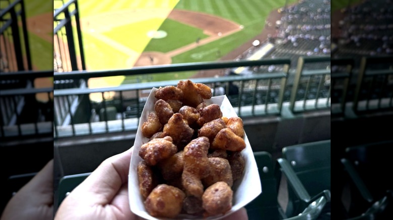 A hand holding a tray of fried cheese curds, in the stands above a baseball diamond