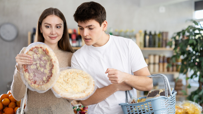 a woman holding two frozen pizzas and a man holding a grocery basket try to decide between the pizzas
