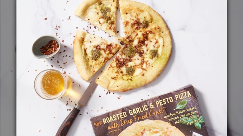 a Trader Joe's roasted garlic and pesto pizza next to ramekins of oil and red pepper flakes and a box of the product on a white board