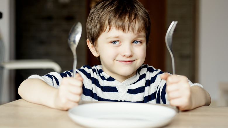 A child sitting with fork and spoon at the table with empty plate