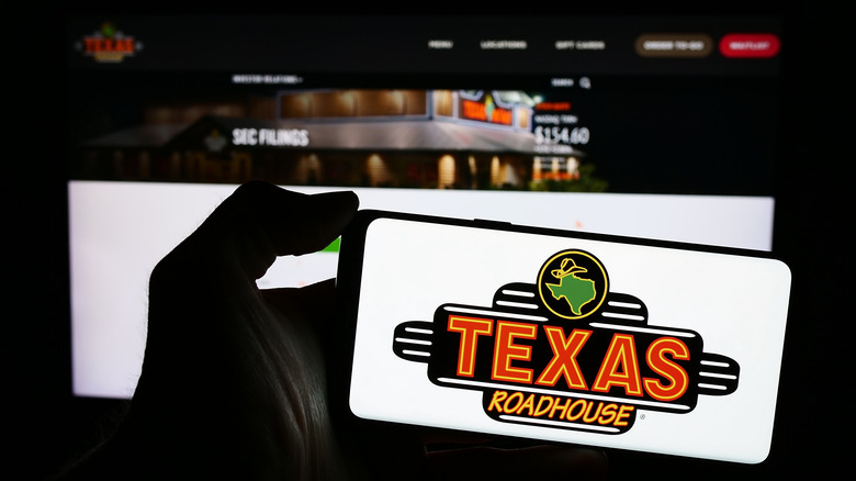 A person holds up a phone with the Texas Roadhouse logo on it in front of a computer screen