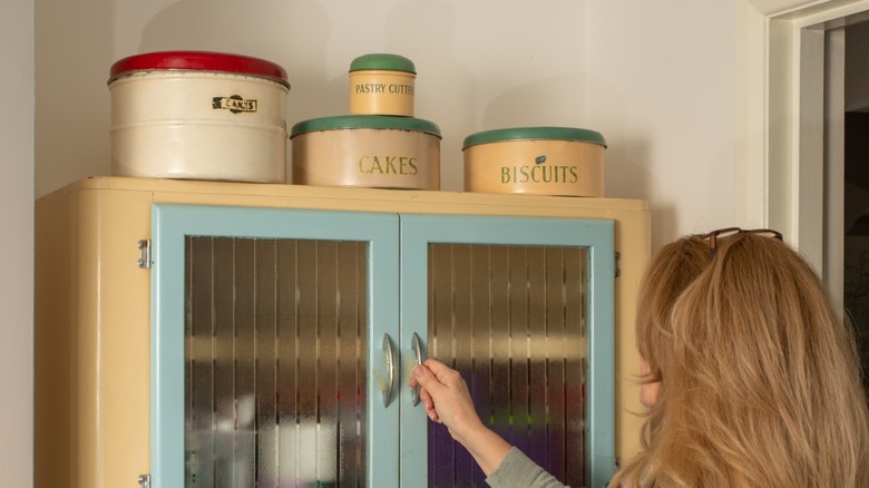 A woman opening a cabinet with antique tins on top