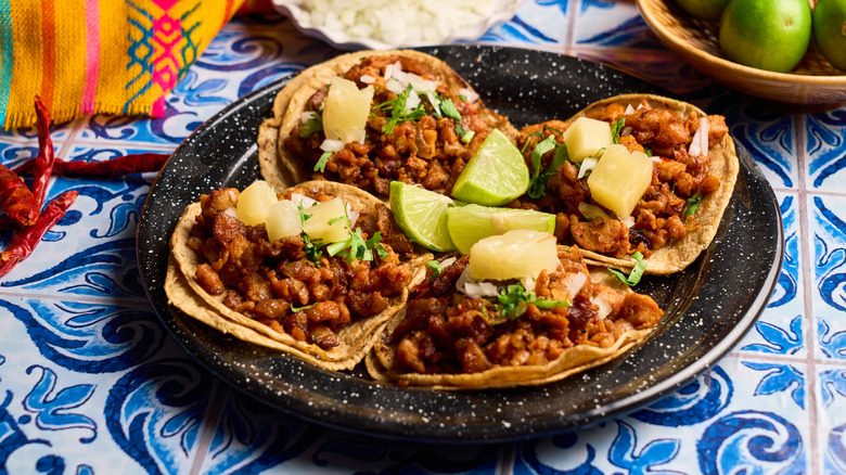 Four street tacos al pastor on a black plate with lime wedges