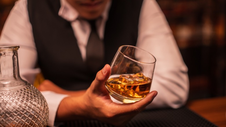 A close up of a person holding a glass of whiskey on the rocks next to a round decanter