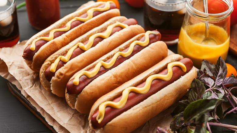 Four hot dogs with mustard and a jar of mustard behind them
