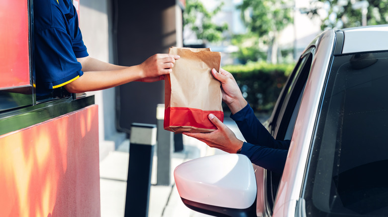 A McDonald's worker hands a customer a bag of food in a drive-thru, with the customer receiving it in their car
