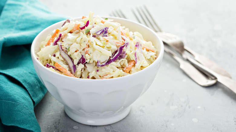 A white bowl filled with coleslaw with a napkin and utensils on the side