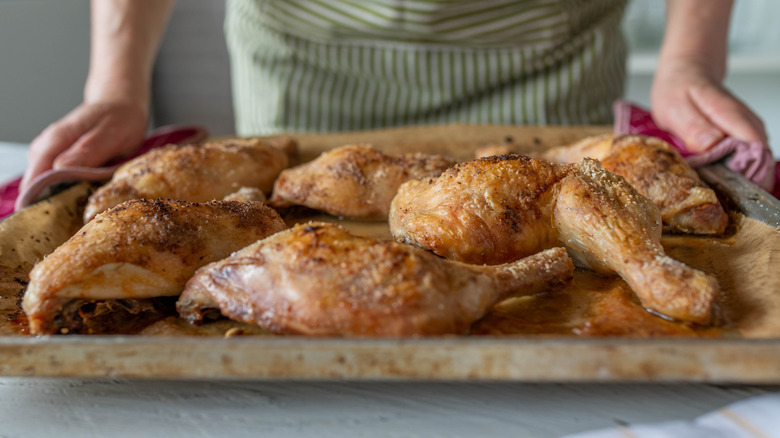 A person is holding a baking sheet with roast chicken legs on it