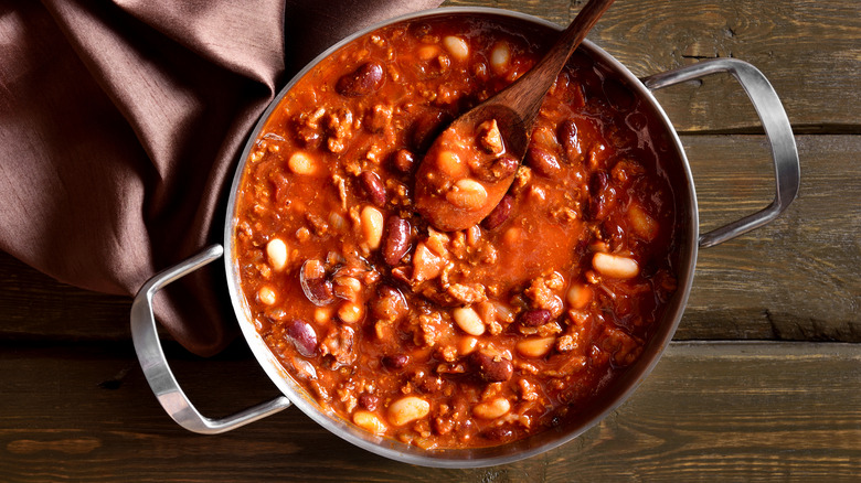 A metal pot of chili with a wooden spoon sits on a table.
