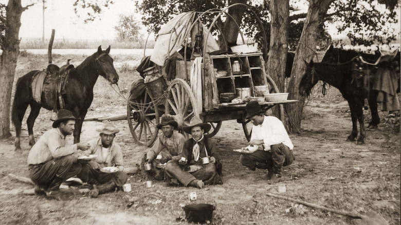 Cowboys sit in front of the chuck wagon in a black and white photograph.
