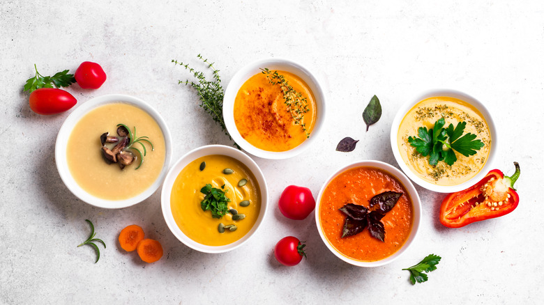 Five autumnal soups in shades of red, yellow, and oranges shot from above on a white table littered with fresh vegetables