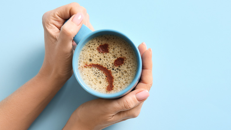 A pair of hands holding a coffee cup with a sad face drawn on the foam