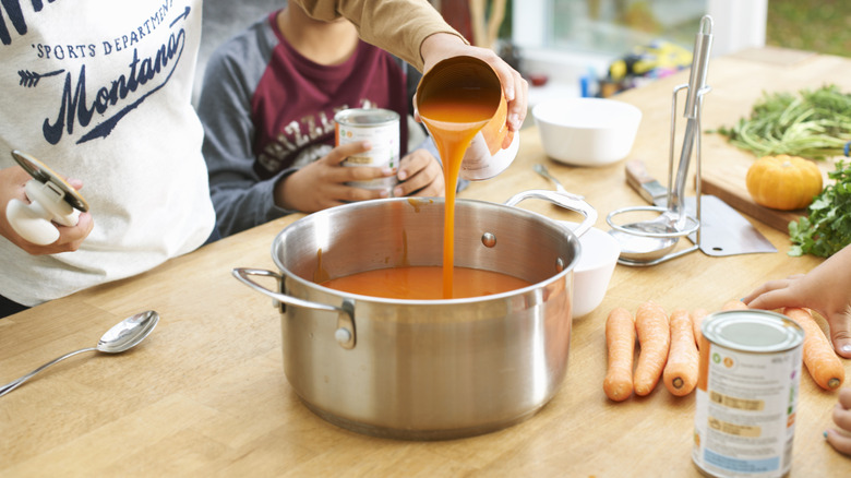 Adult pouring canned tomato soup into a pot with child watching