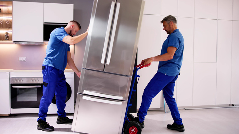 Two guys in uniform moving a refrigerator in a kitchen