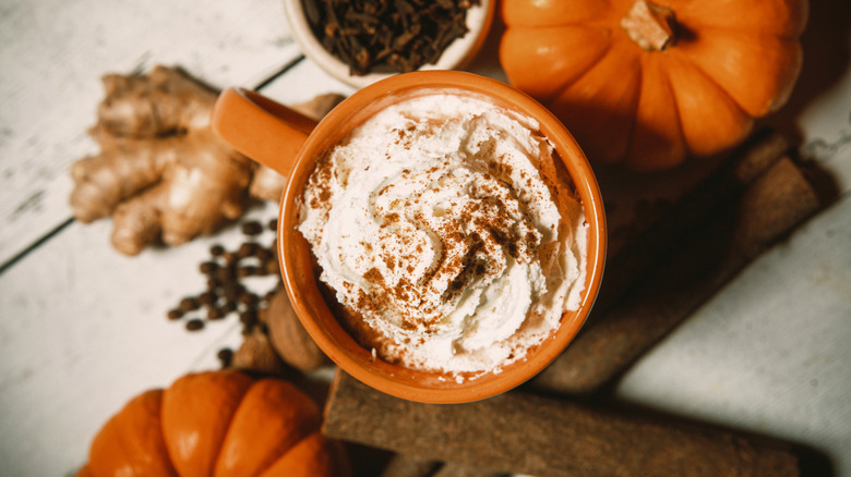 A pumpkin spice latte in an orange mug surrounded by pumpkins and various spices