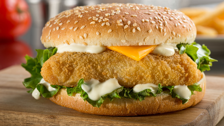 A fried fish sandwich with cheese, lettuce, sauce, and a sesame seed bun on a wood board