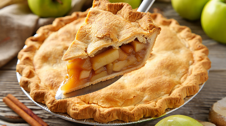A slice of apple pie with the rest of the pie behind it on a table with green apples nearby.