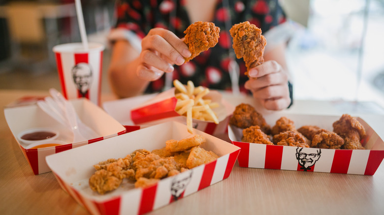 Person holding two KFC fried chicken drumsticks at a table with KFC food
