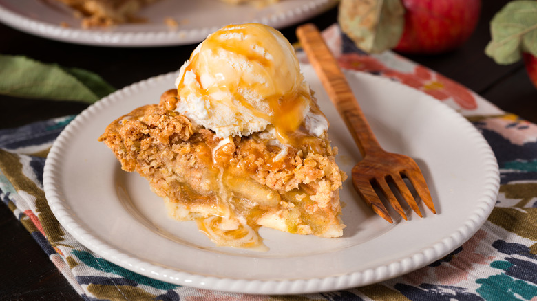 Apple pie with ice cream on top and drizzled with honey on a white plate with a wooden fork next to it.