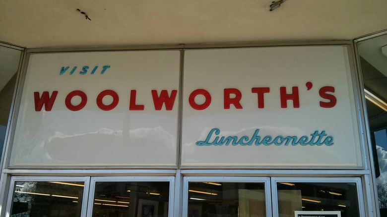 A vintage white sign that says "Visit Woolworth's Luncheonette" in alternating light blue and red text