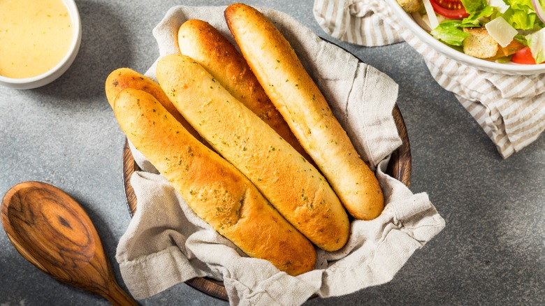Basket of fresh breadsticks on a table next to a wooden spoon and a bowl of salad