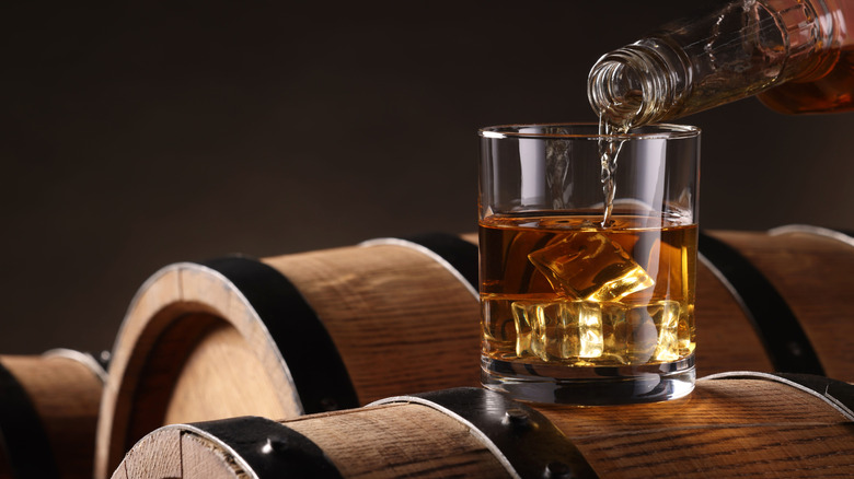 Person pouring whiskey into glass on barrel