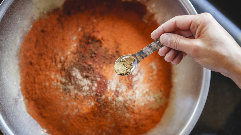 A large bowl of brightly colored spice mix shot from above as a hand adds a spice