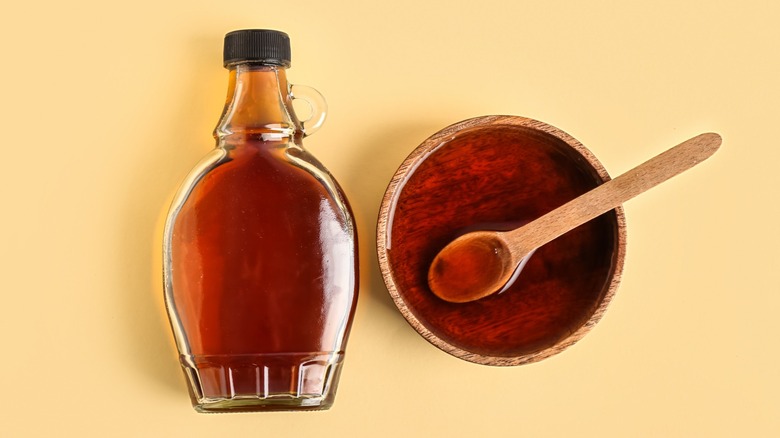 A bottle of maple syrup with small handle and scalloped bottom. A bowl of syrup is next to it