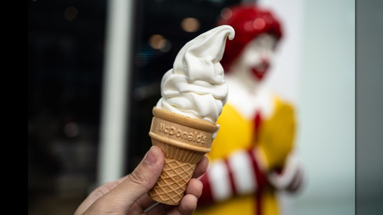 A hand holding a McDonald's vanilla ice cream cone in front of a Ronald McDonald statue