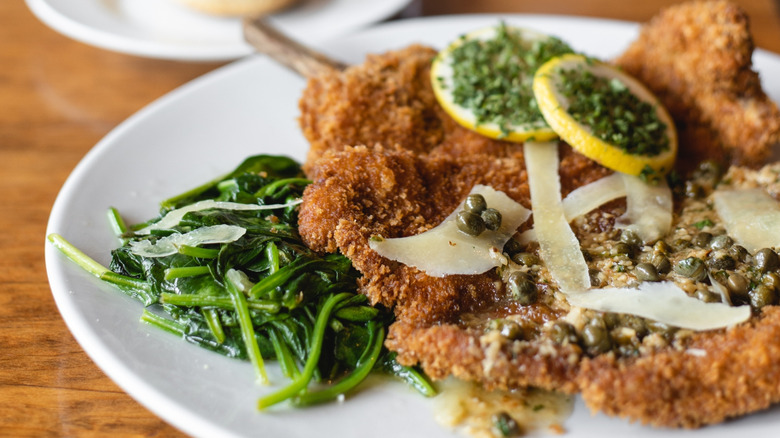 A plate of veal Milanese with lemons and parmesan cheese on it and greens beside it
