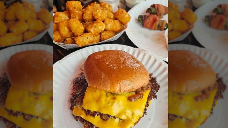A photo of a smash burger with cheese on a paper plate next to a pile of tater tots
