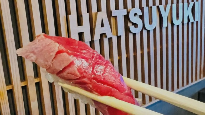 A piece of sashimi being held up by chopsticks in front of a wooden sign saying Hatsuyuki