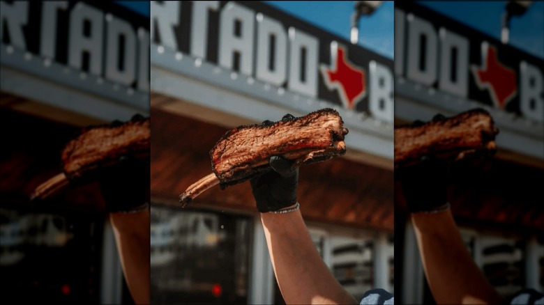 A hand holds a meaty rin in front of Hurtado's BBq sign