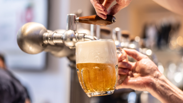 A close-up of a person's hand holding a pint glass at an angle being filled with lager. The lager is golden in color creating foam and bubbles.