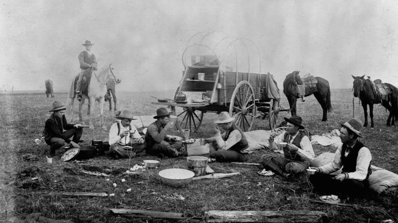 Black and white photo of cowboys having meal in front of wagon and horses