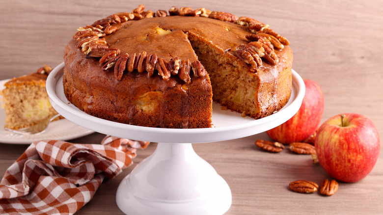 Spiced apple cider cake with pecans on a white cake stand with apples and a gingham napkin on the table.