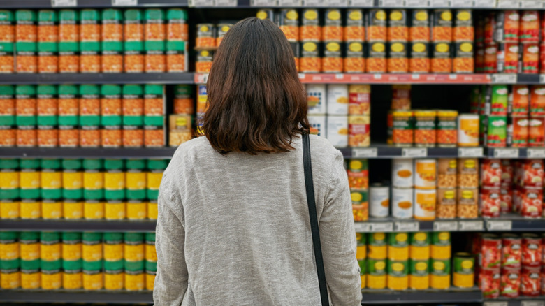 A woman stands in front of a supermarket shelf filled with out of focus canned products