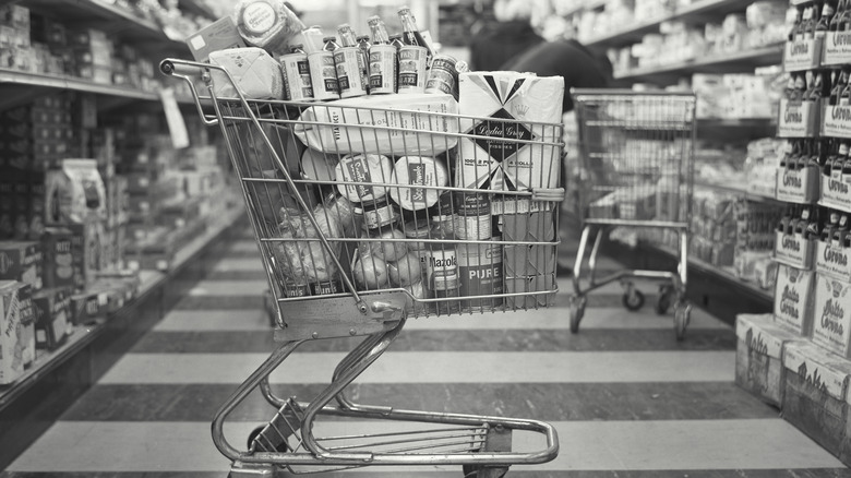 A black and white photo of a vintage grocery cart full of products in an aisle