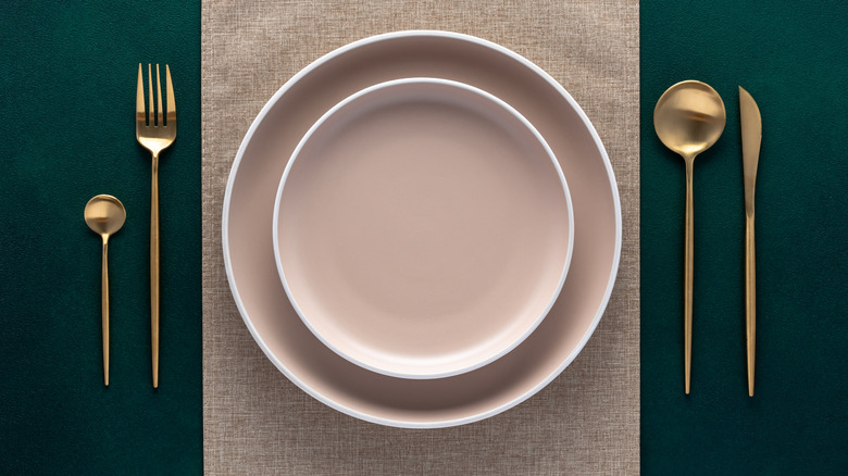 A table setting with white plates on a beige place setting, and gold cutlery on either side on a green table