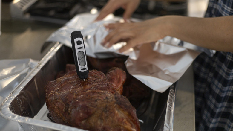 Meat thermometer in prime rib as it sits in a tray being covered with foil by two hands
