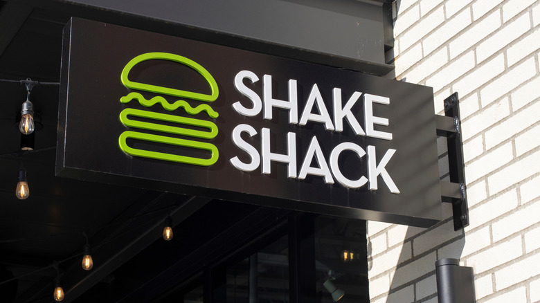 A black Shake Shack sign hanging on a white brick wall with a lightbulb beside it.