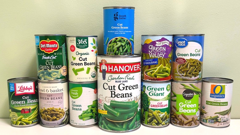 Twelve cans of green beans stacked up on a table against a wall