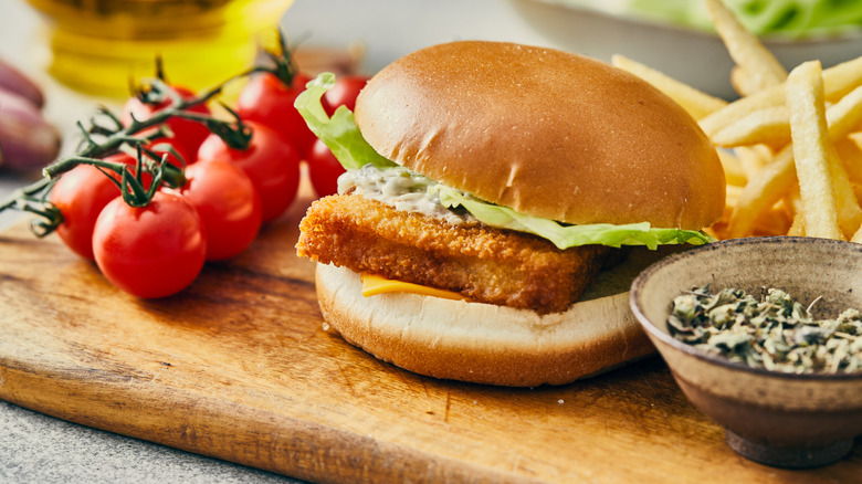 A fish burger on a bread board, surrounded by ingredients