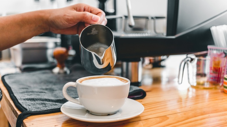 A cappuccino in a white porcelain cup with foam poured from a silver frothing pitcher