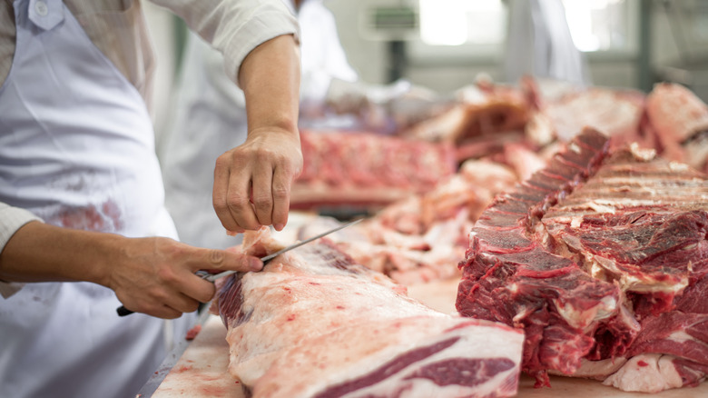 Butcher slicing fat off a joint of meat, with meat cuts around him